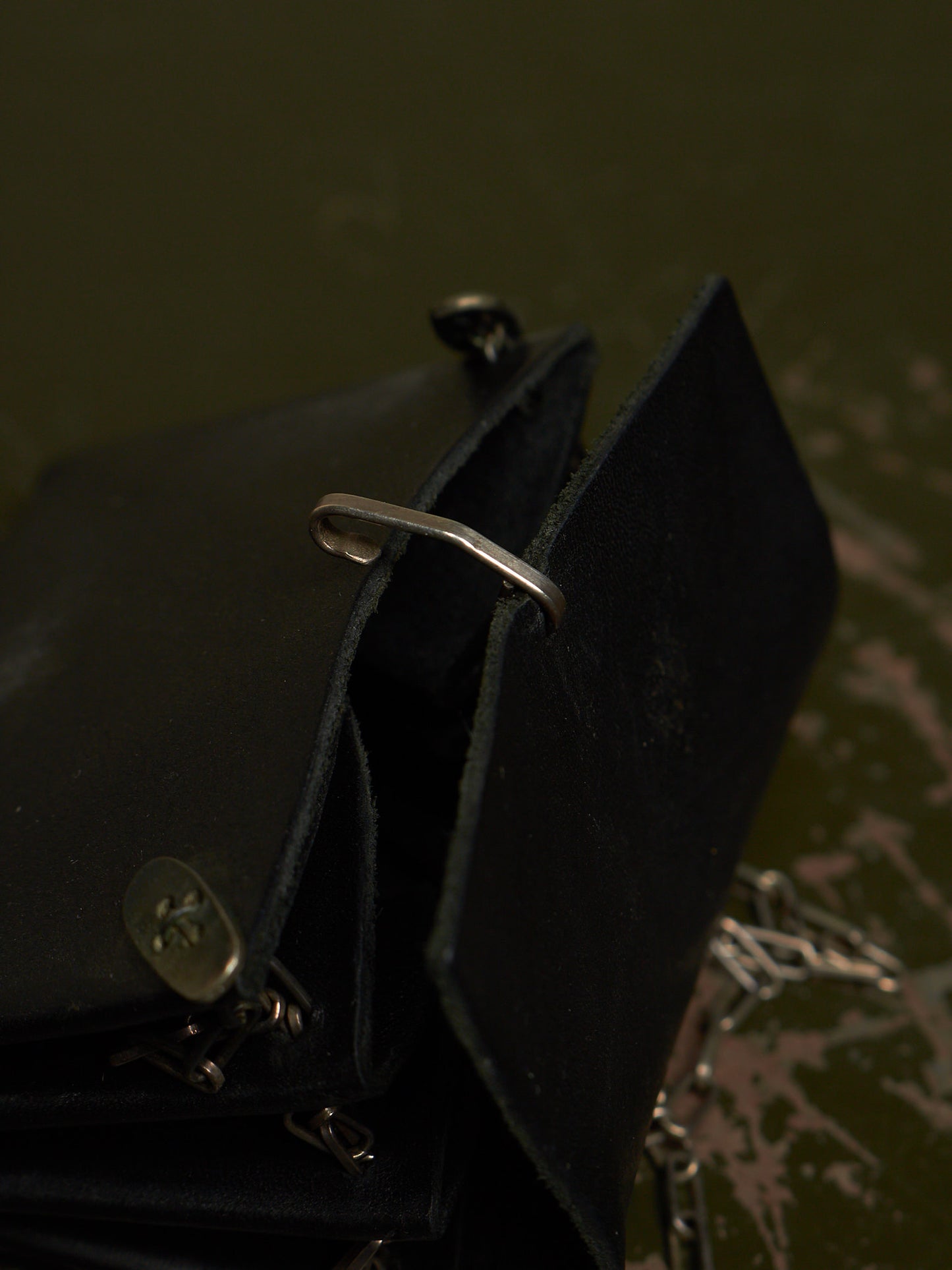A close-up of a black leather wallet or clutch with a silver-tone metal ring detail and chain, set against a dark olive green background.
