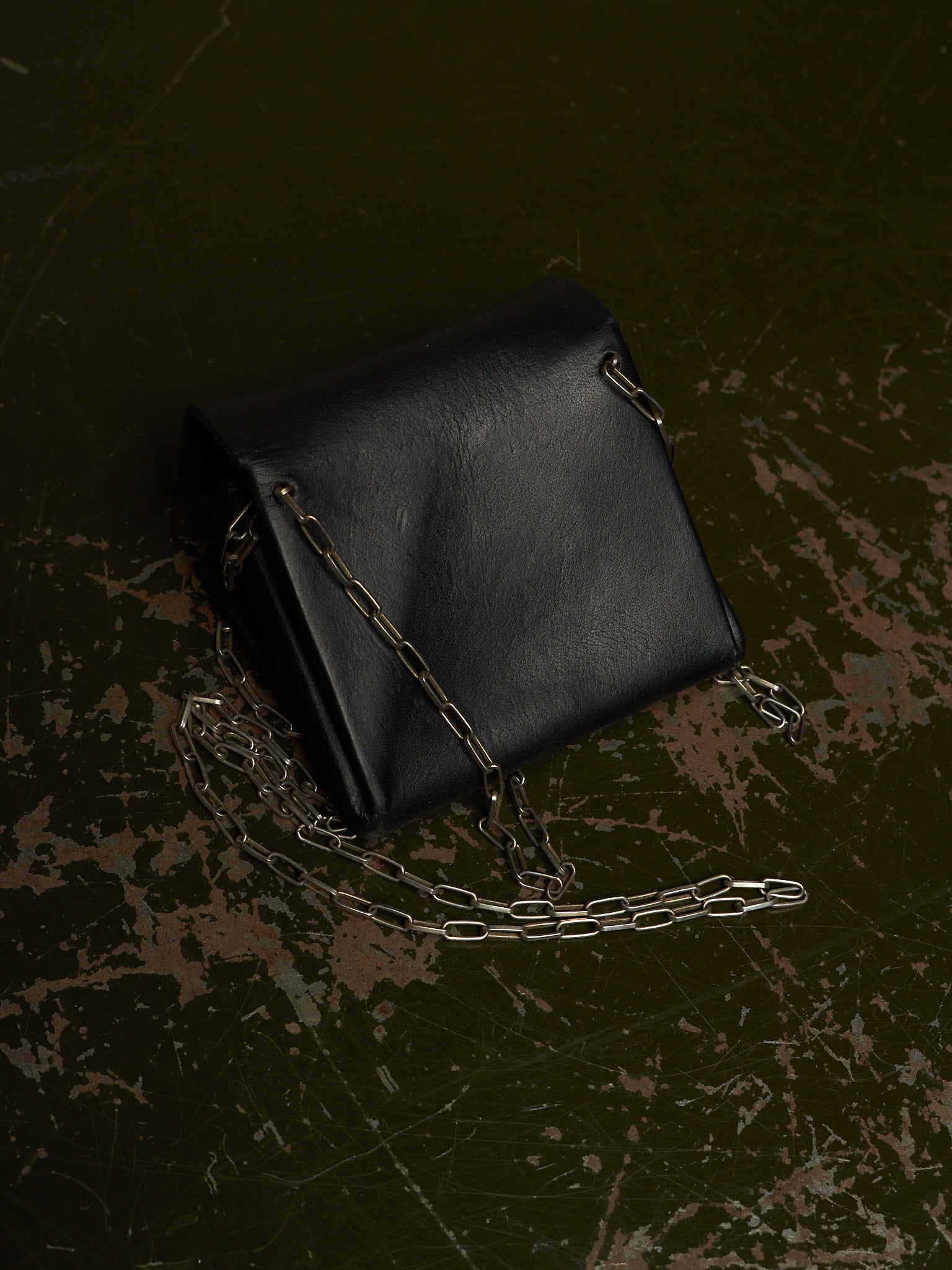 A black leather shoulder bag with a silver chain strap, photographed from an overhead angle on a dark, distressed textured surface.