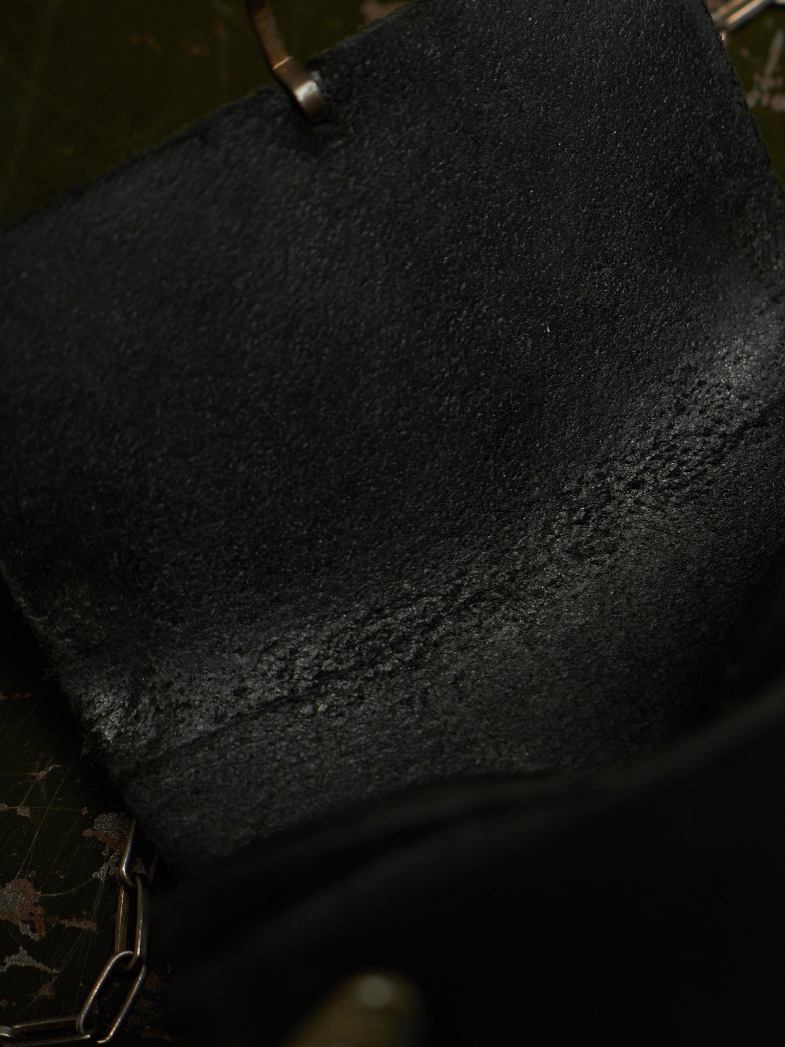 Close-up of a black textured leather or suede material with a small metal rivet and chain detail visible at the top and bottom edges.