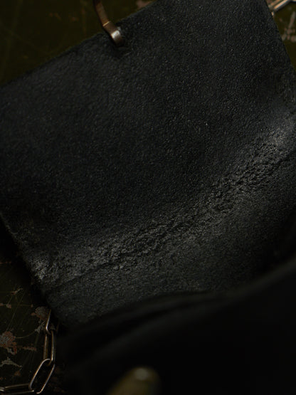 Close-up of a black textured leather or suede material with a small metal rivet and chain detail visible at the top and bottom edges.