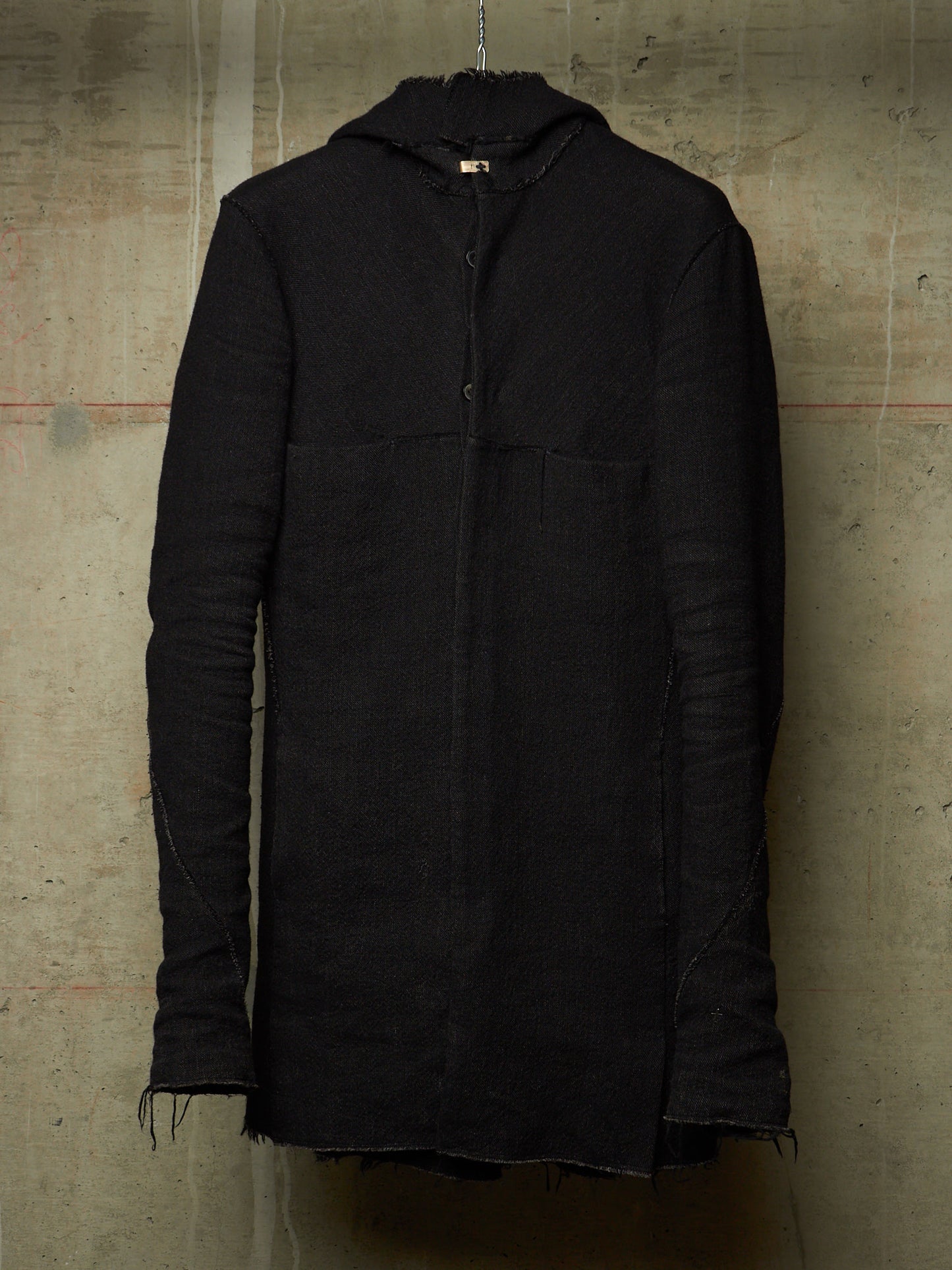 A black hooded jacket with a textured, ribbed fabric and raw frayed edges, hanging against a concrete wall.