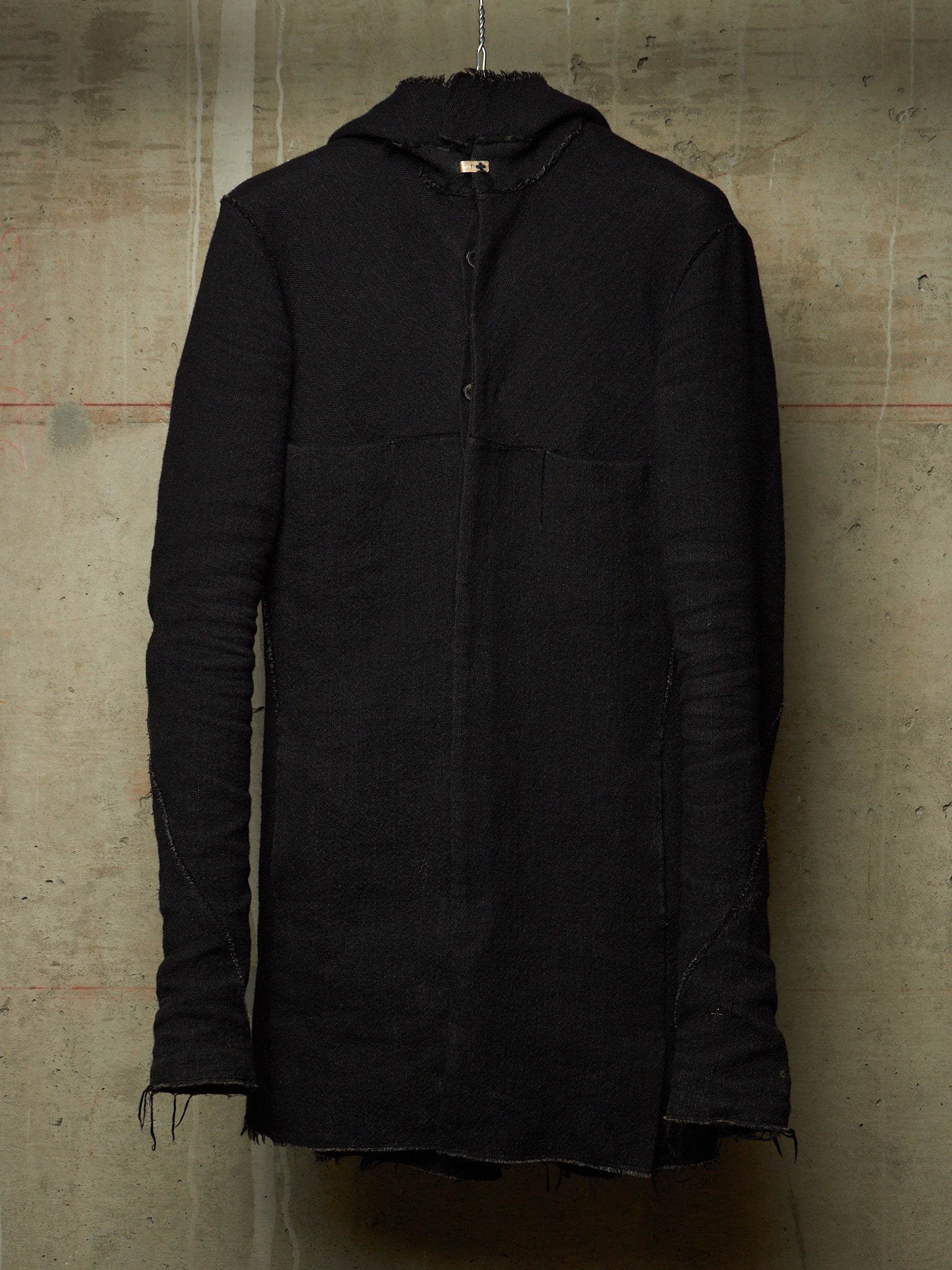 A black hooded jacket with a textured, ribbed fabric and raw frayed edges, hanging against a concrete wall.