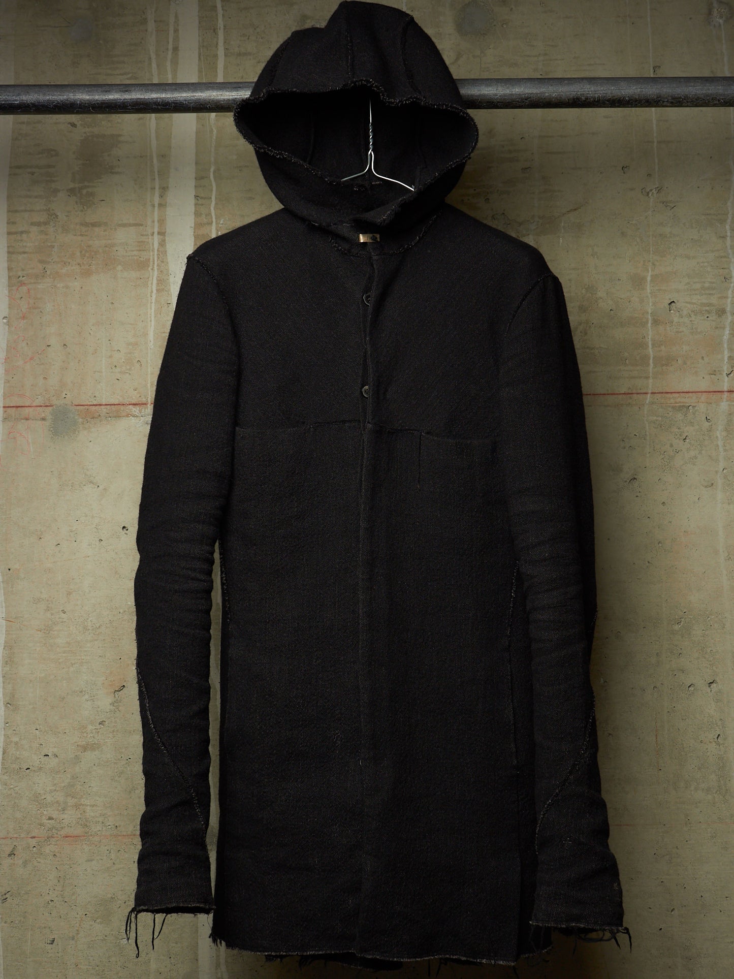 A long black hooded jacket with a button front and raw frayed edges, hanging against a textured concrete wall.