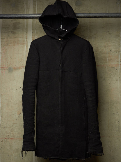 A long black hooded jacket with a button front and raw frayed edges, hanging against a textured concrete wall.