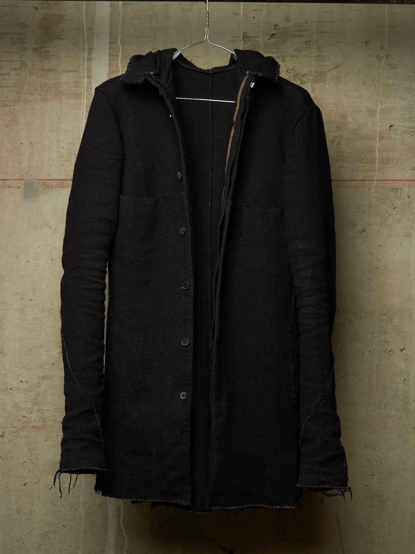 A black hooded button-front coat with raw, frayed edges and front flap pockets, hanging on a wire hanger against a textured concrete wall.