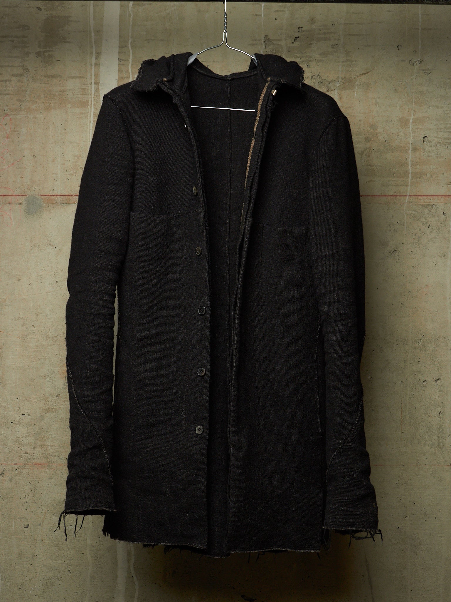 A black hooded button-front coat with raw, frayed edges and front flap pockets, hanging on a wire hanger against a textured concrete wall.