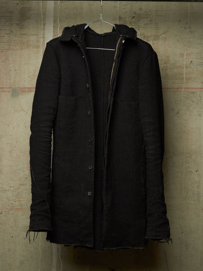 A black hooded button-front coat with raw, frayed edges and front flap pockets, hanging on a wire hanger against a textured concrete wall.
