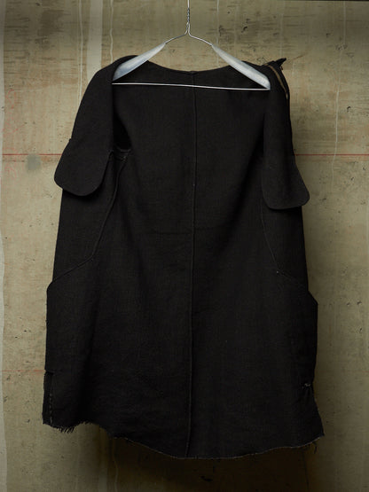A black linen-blend tunic top with wide shoulder cutouts and a central seam, hanging on a metal hanger against a textured concrete wall.