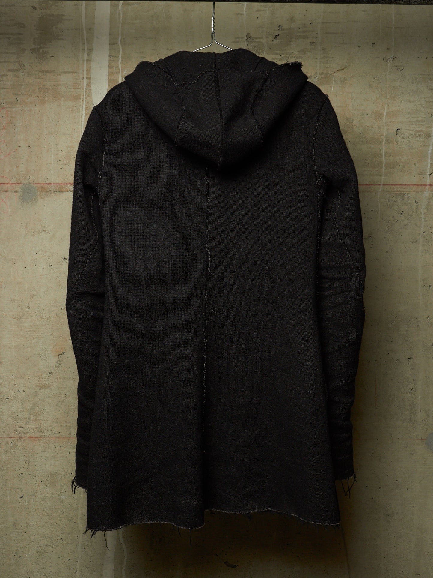 A black hooded jacket with raw, frayed edges and distressed detailing, hanging on a wire hanger against a textured concrete wall.