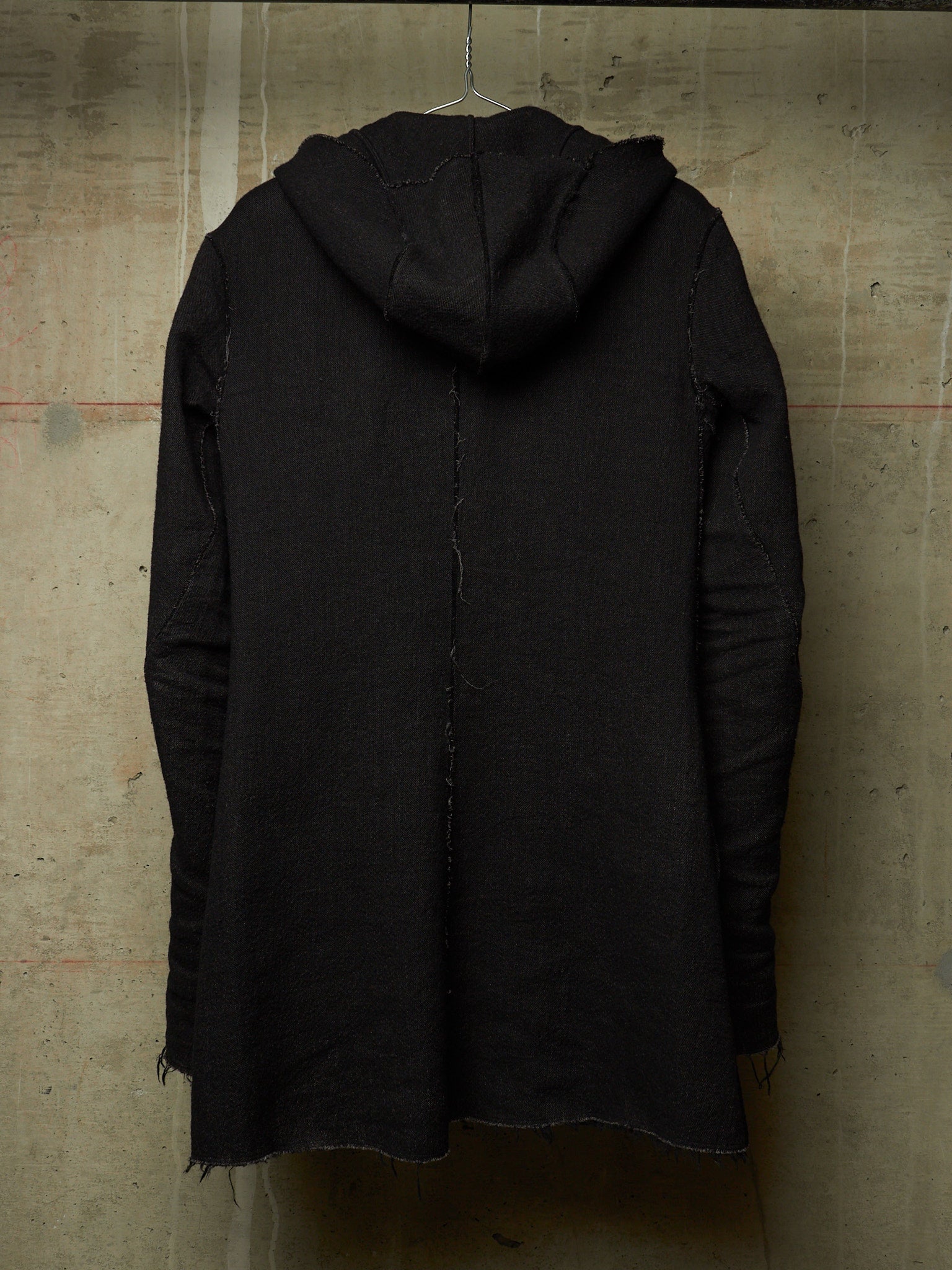 A black hooded jacket with raw, frayed edges and distressed detailing, hanging on a wire hanger against a textured concrete wall.