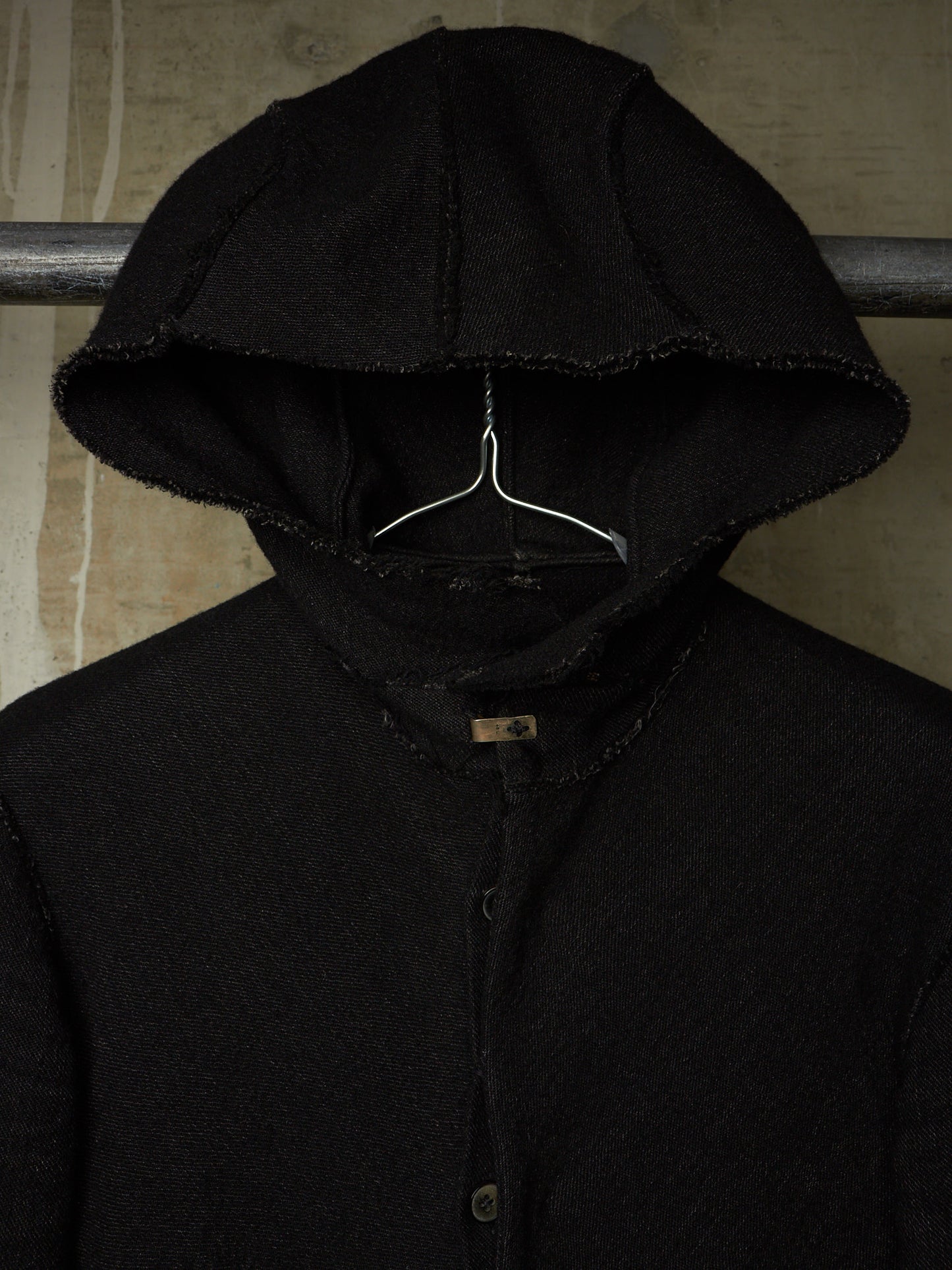 A black hooded jacket with raw, frayed edges hanging on a metal hanger against a textured beige wall.