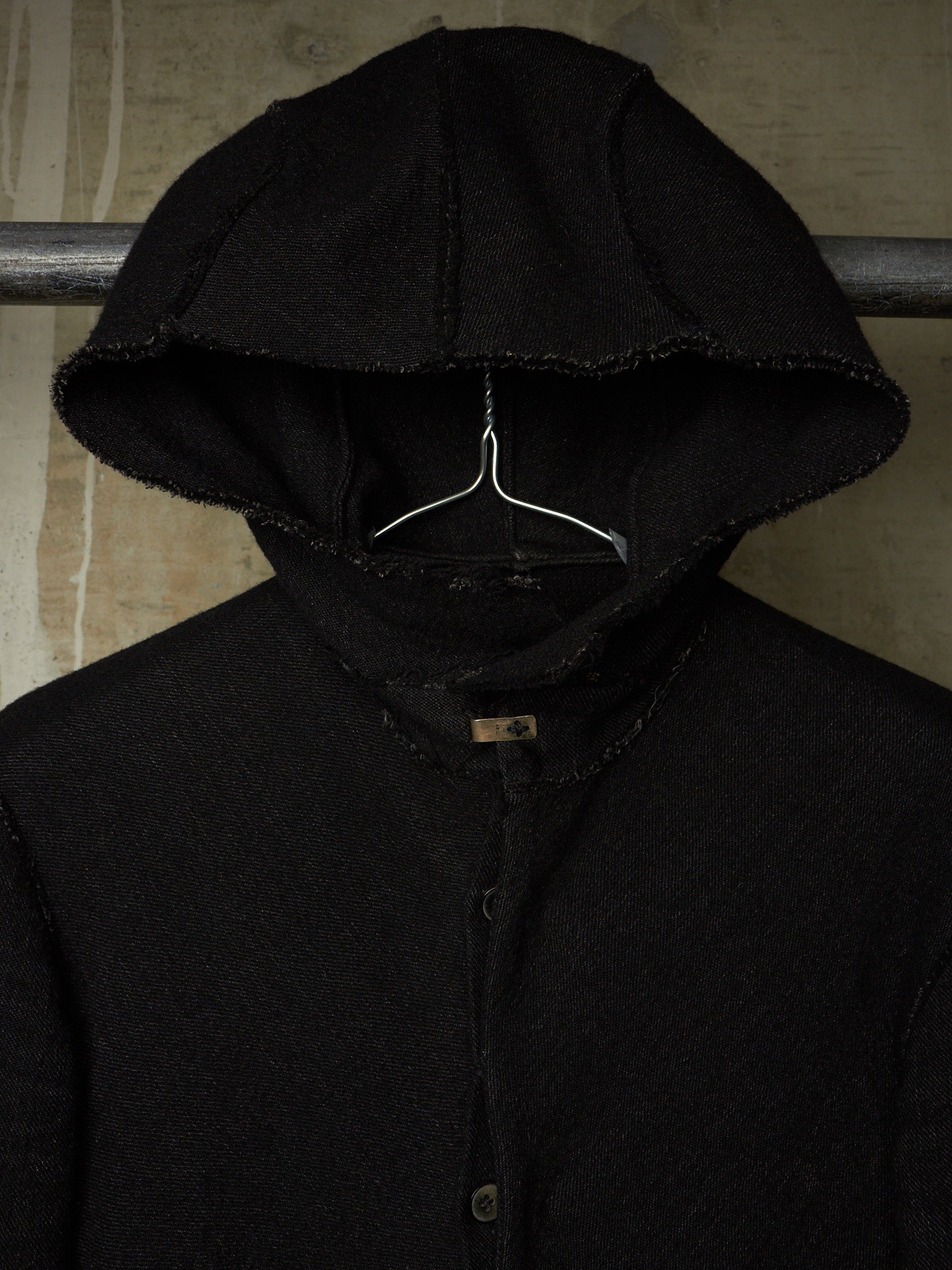 A black hooded jacket with raw, frayed edges hanging on a metal hanger against a textured beige wall.