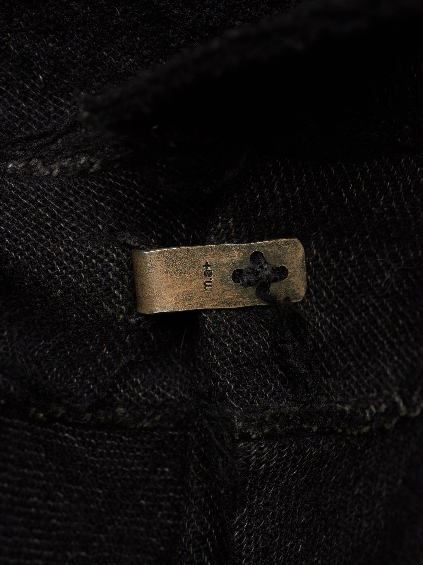 Close-up of a black textured fabric garment with a small rectangular brass button featuring a clover-like cutout and the brand name 'KNU' stitched vertically.