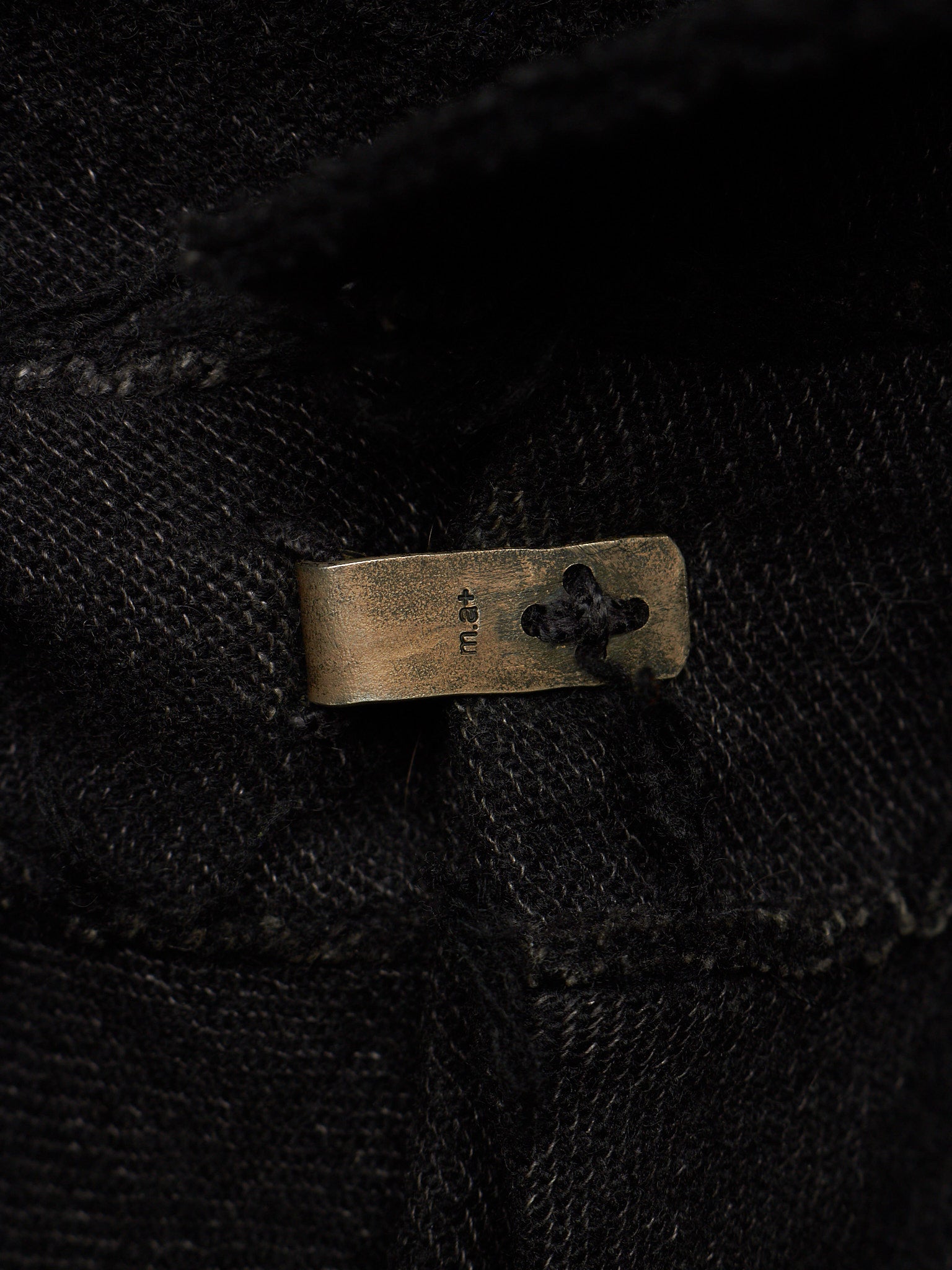 Close-up of a black textured fabric garment with a small rectangular brass button featuring a clover-like cutout and the brand name 'KNU' stitched vertically.