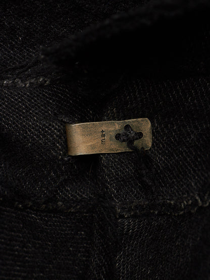 Close-up of a black textured fabric garment with a small rectangular brass button featuring a clover-like cutout and the brand name 'KNU' stitched vertically.