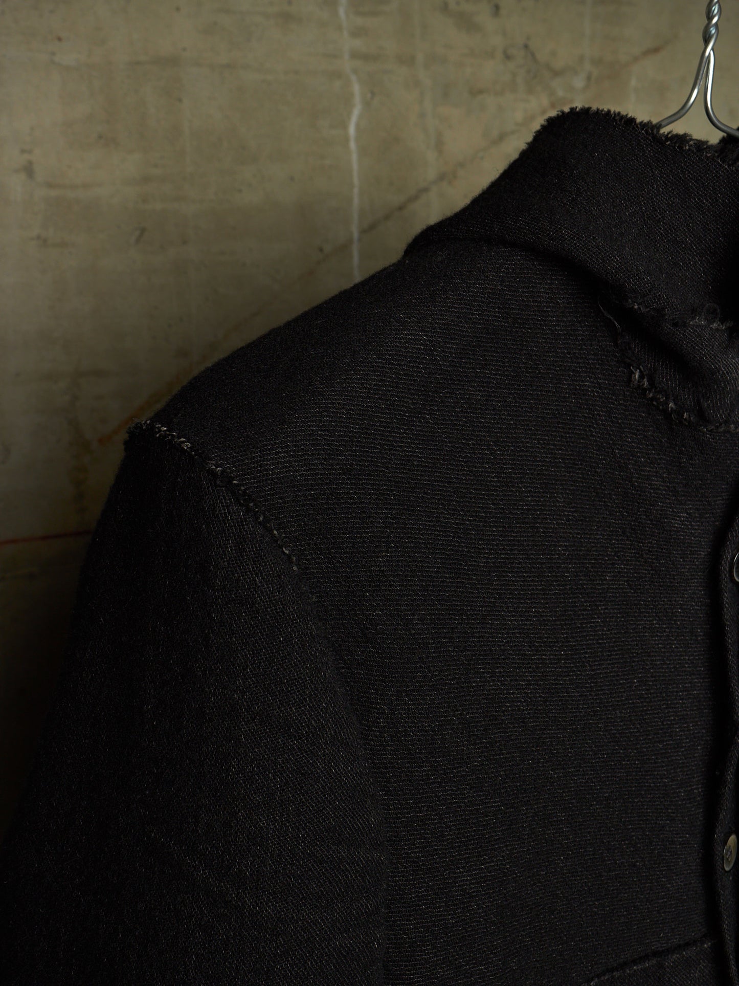 Close-up of a black textured jacket or overshirt on a hanger, showing the collar, shoulder seam, and fabric weave against a warm beige wall background.