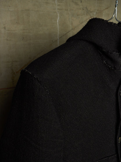Close-up of a black textured jacket or overshirt on a hanger, showing the collar, shoulder seam, and fabric weave against a warm beige wall background.