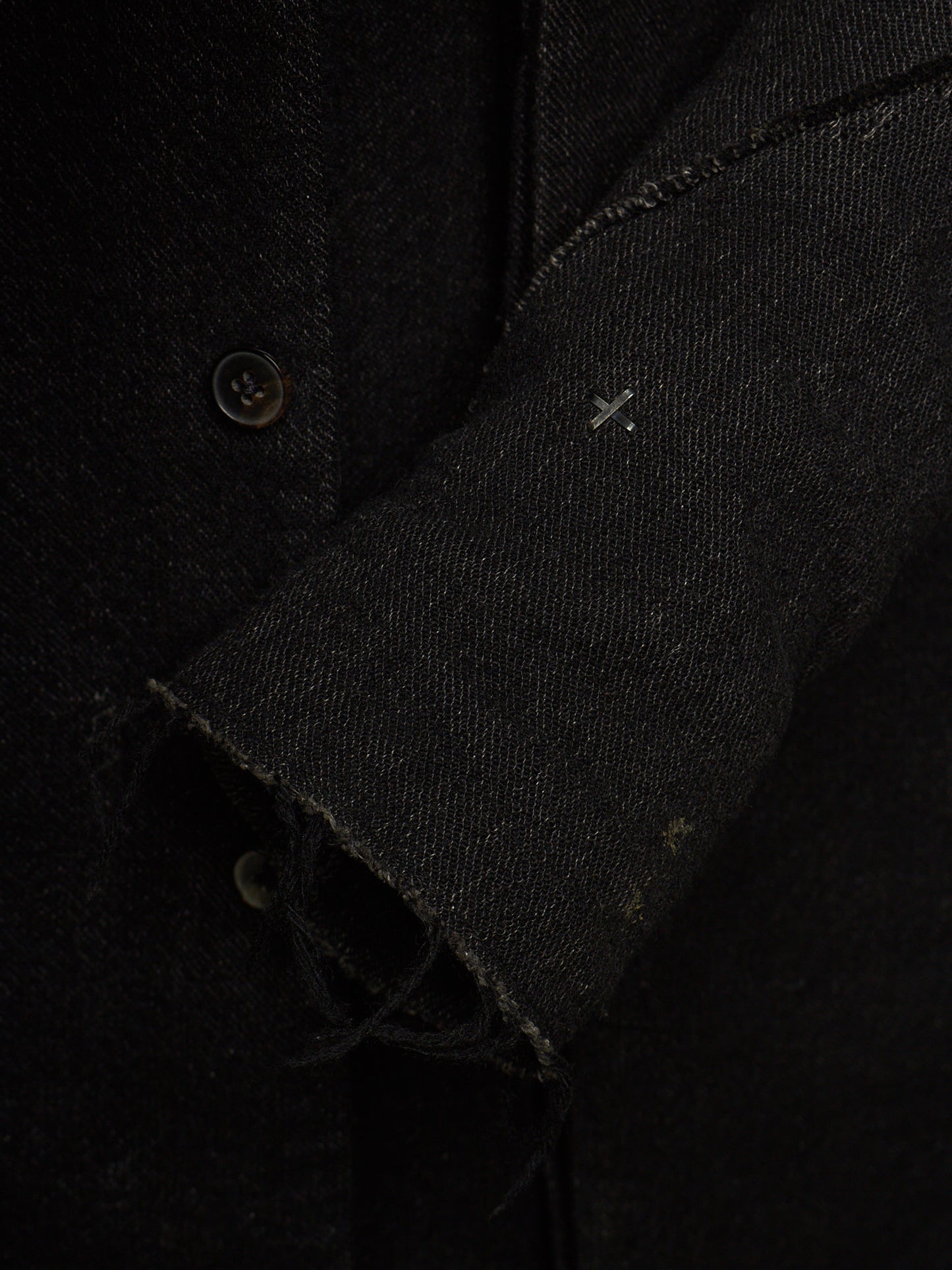 Close-up detail of a black textured fabric garment, showing a buttoned front, raw frayed cuff, and a small white stitched cross detail on the sleeve.