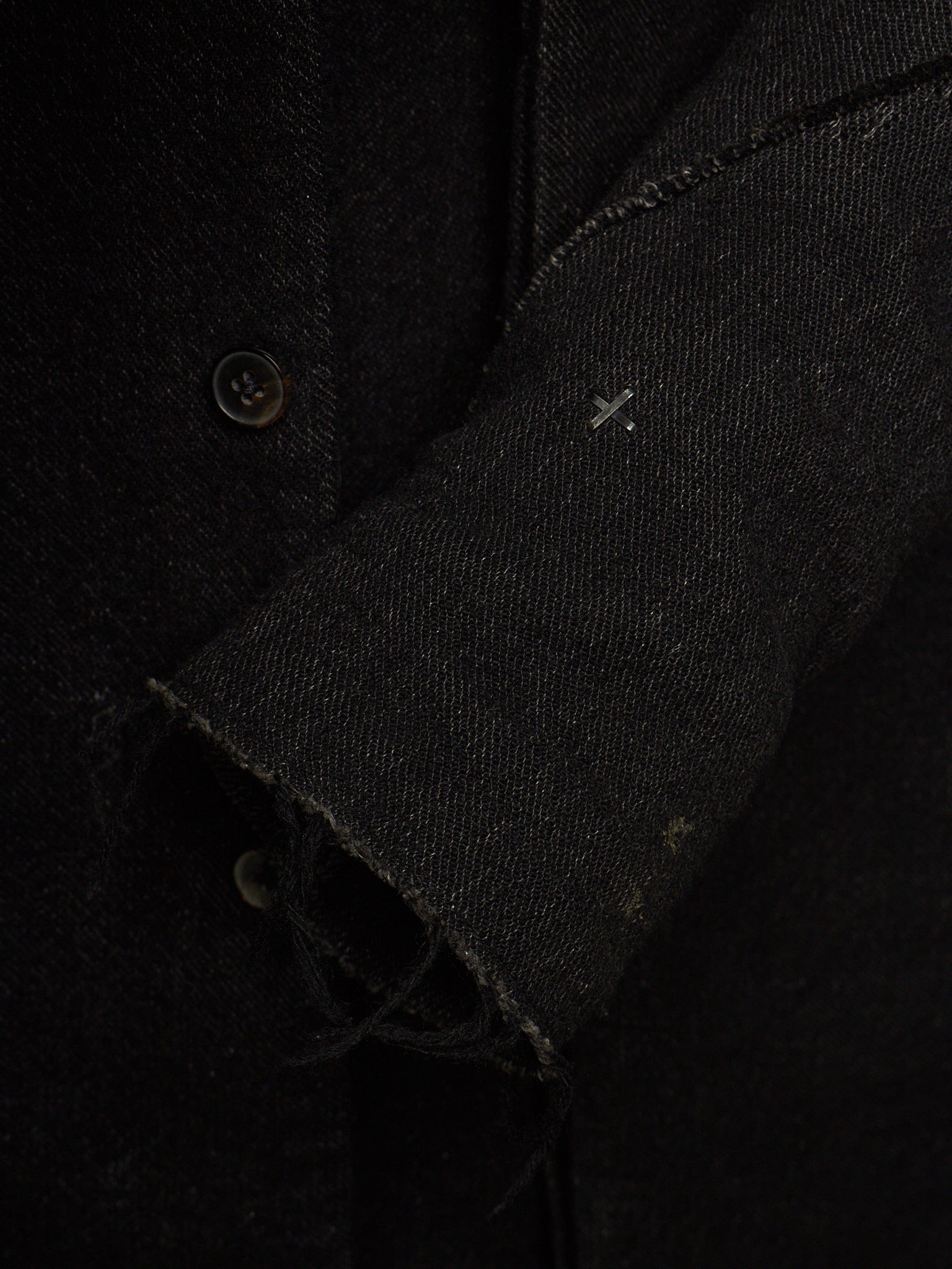 Close-up detail of a black textured fabric garment, showing a buttoned front, raw frayed cuff, and a small white stitched cross detail on the sleeve.