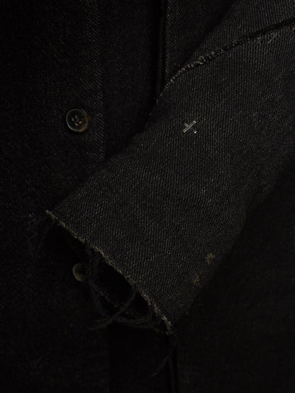 Close-up detail of a black textured fabric garment, showing a buttoned front, raw frayed cuff, and a small white stitched cross detail on the sleeve.