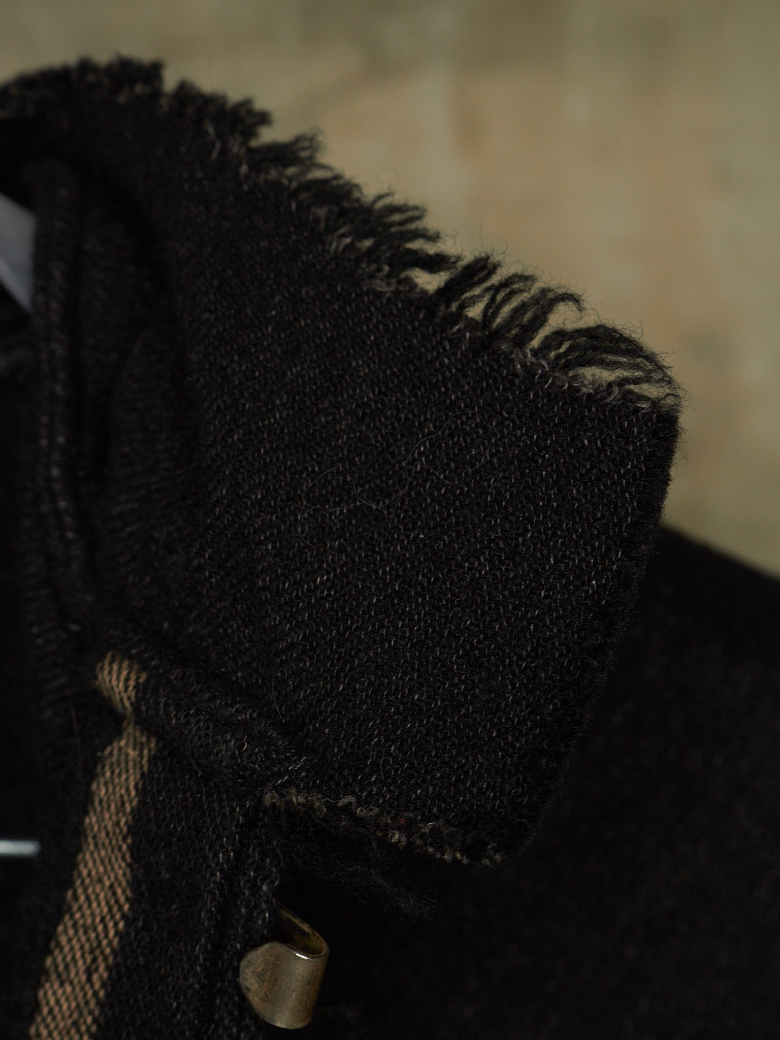 Close-up detail of a black textured fabric garment, showing a frayed collar edge and a small metallic snap button with a tan woven strap.