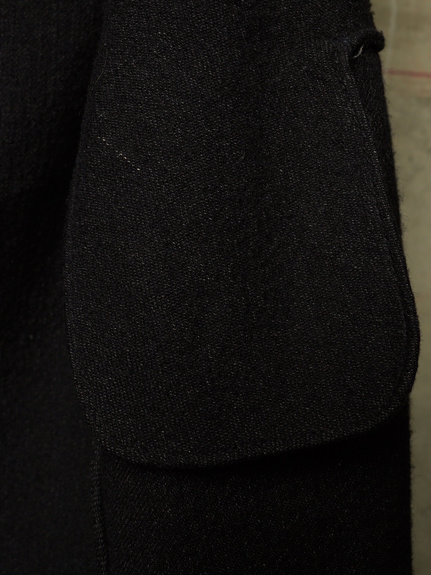 Close-up detail of a black textured fabric garment, showing a large patch pocket and the fine woven texture of the material.