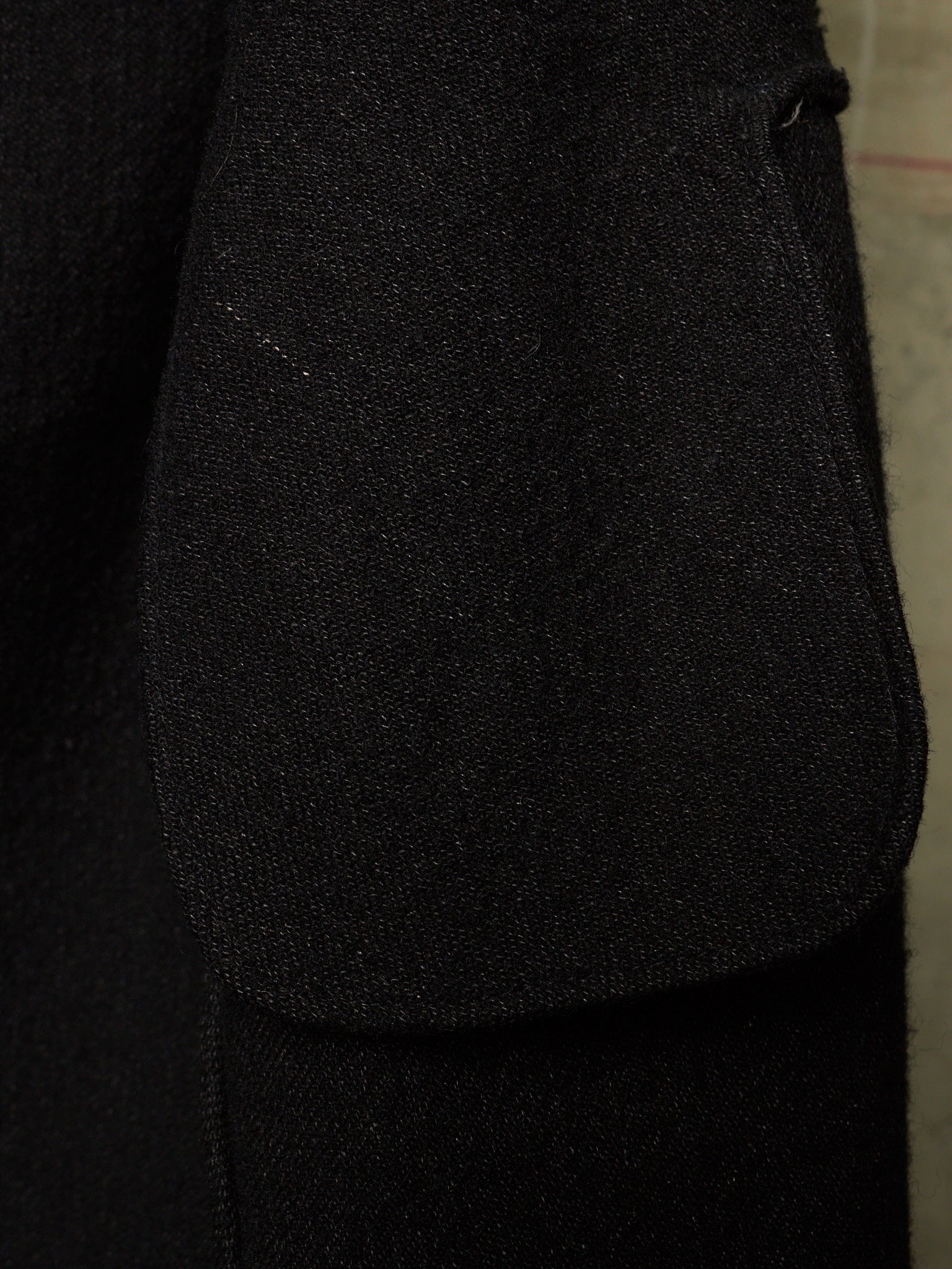 Close-up detail of a black textured fabric garment, showing a large patch pocket and the fine woven texture of the material.