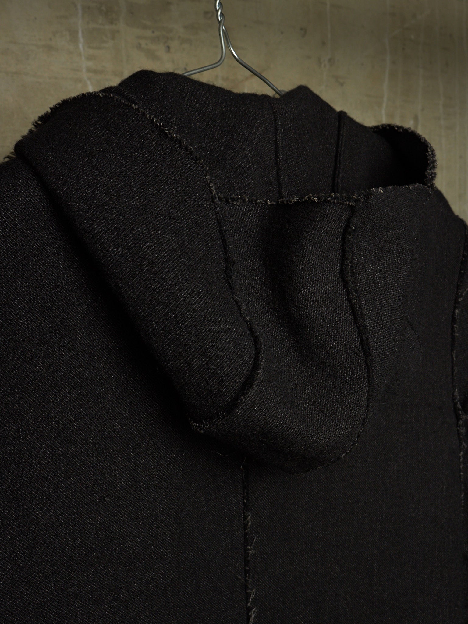 Close-up of a black hooded garment hanging on a metal hanger against a textured beige wall.
