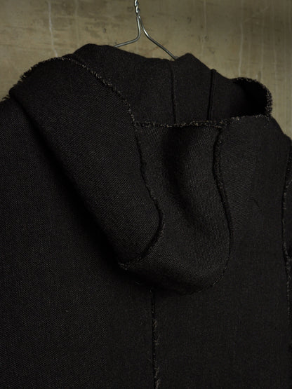 Close-up of a black hooded garment hanging on a metal hanger against a textured beige wall.