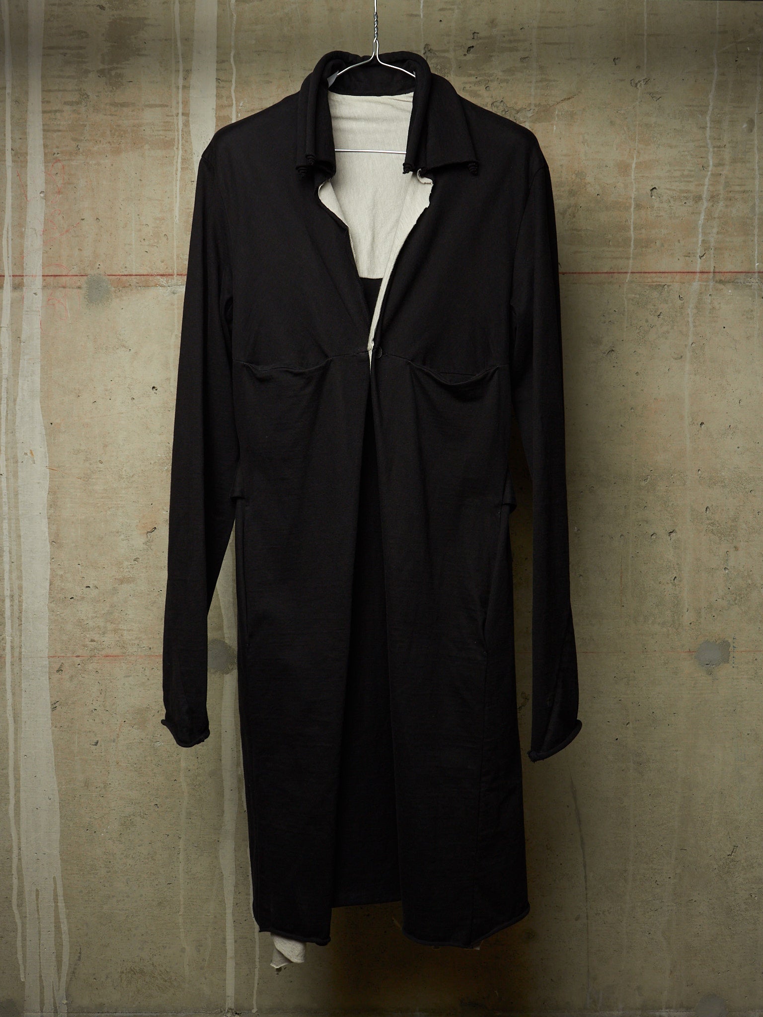 A black long-sleeve collared coat or overshirt hanging on a black hanger against a textured concrete wall, layered over a white top.