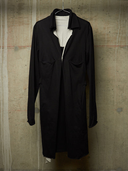A black long-sleeve collared coat or overshirt hanging on a black hanger against a textured concrete wall, layered over a white top.