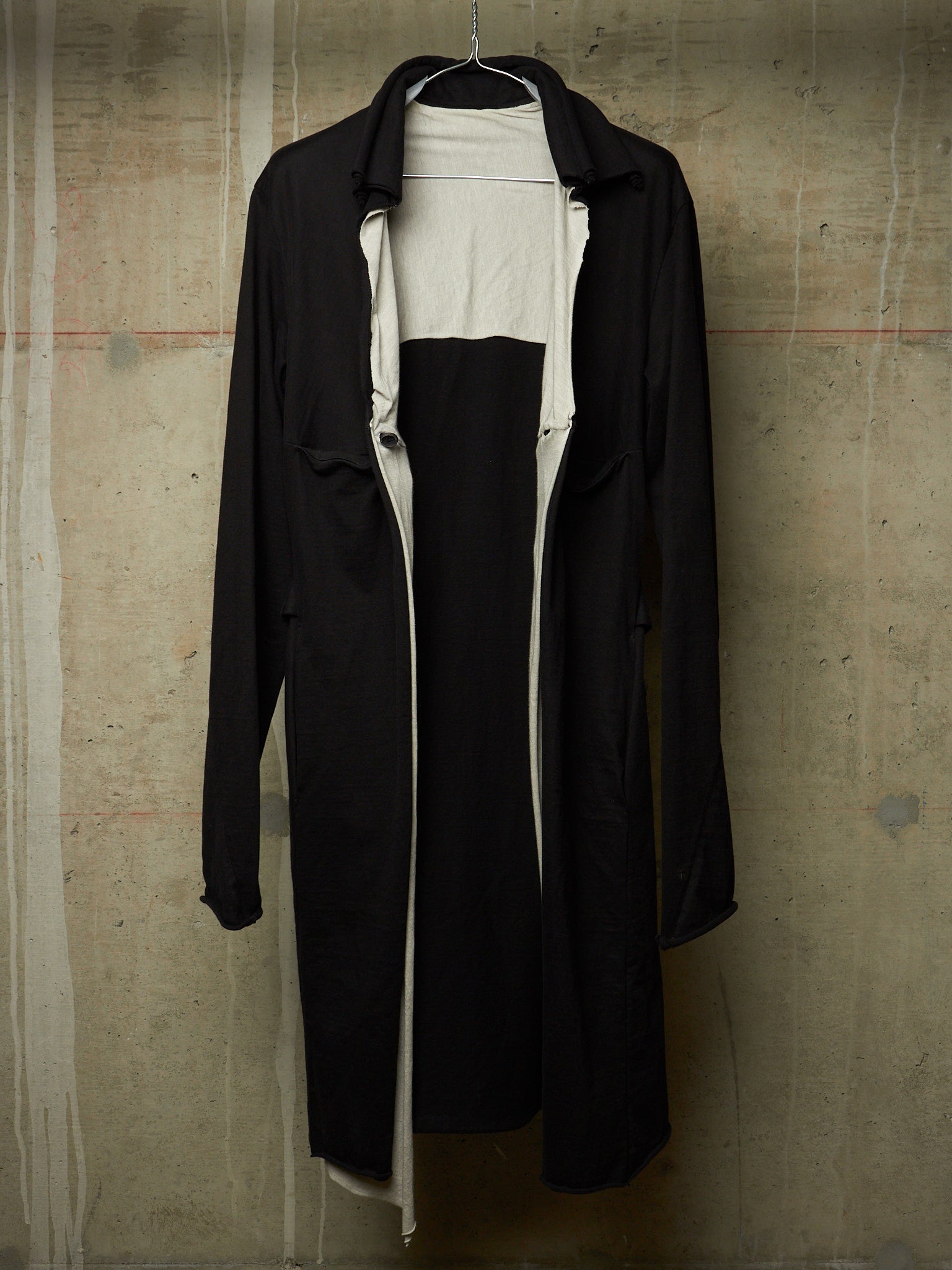A black longline trench coat with a wide collar, hanging on a wire hanger against a textured concrete wall. The coat is open, revealing a white inner layer or dress underneath.