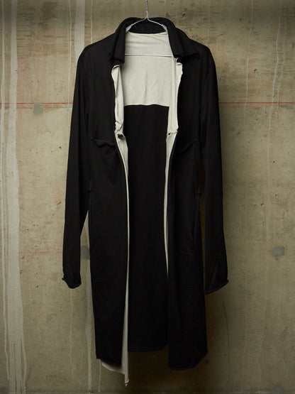 A black longline trench coat with a wide collar, hanging on a wire hanger against a textured concrete wall. The coat is open, revealing a white inner layer or dress underneath.