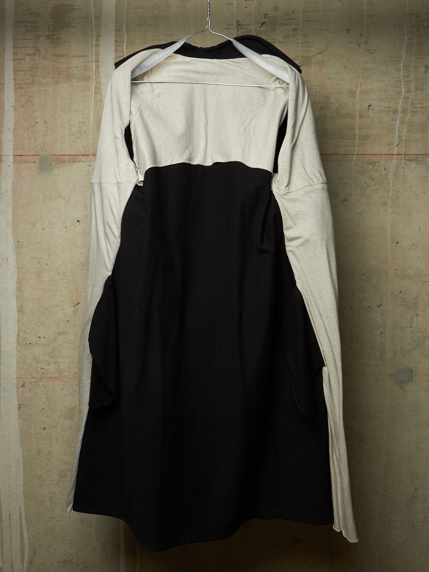 A black and off-white long-sleeve dress with a structured black bodice and flared skirt, hanging on a wire hanger against a textured concrete wall.