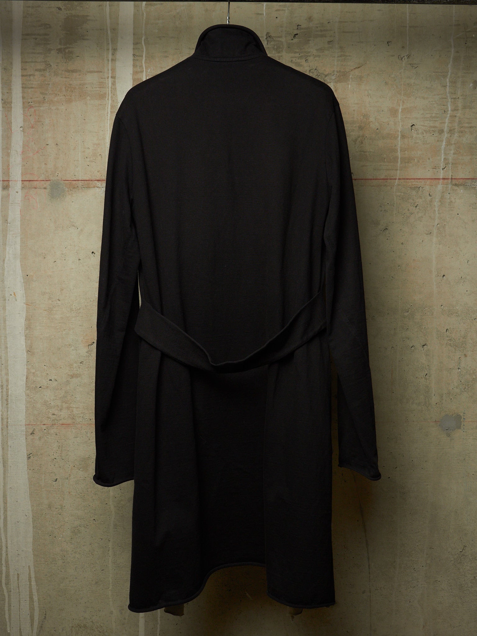 A black long-sleeve turtleneck coat with a self-tie belt, shown from the back against a textured concrete wall.