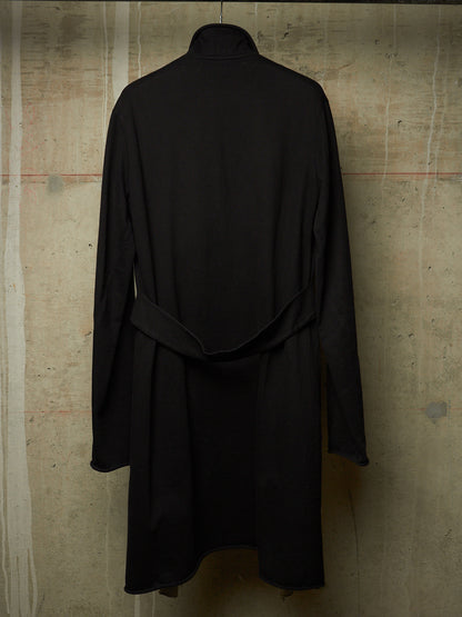 A black long-sleeve turtleneck coat with a self-tie belt, shown from the back against a textured concrete wall.