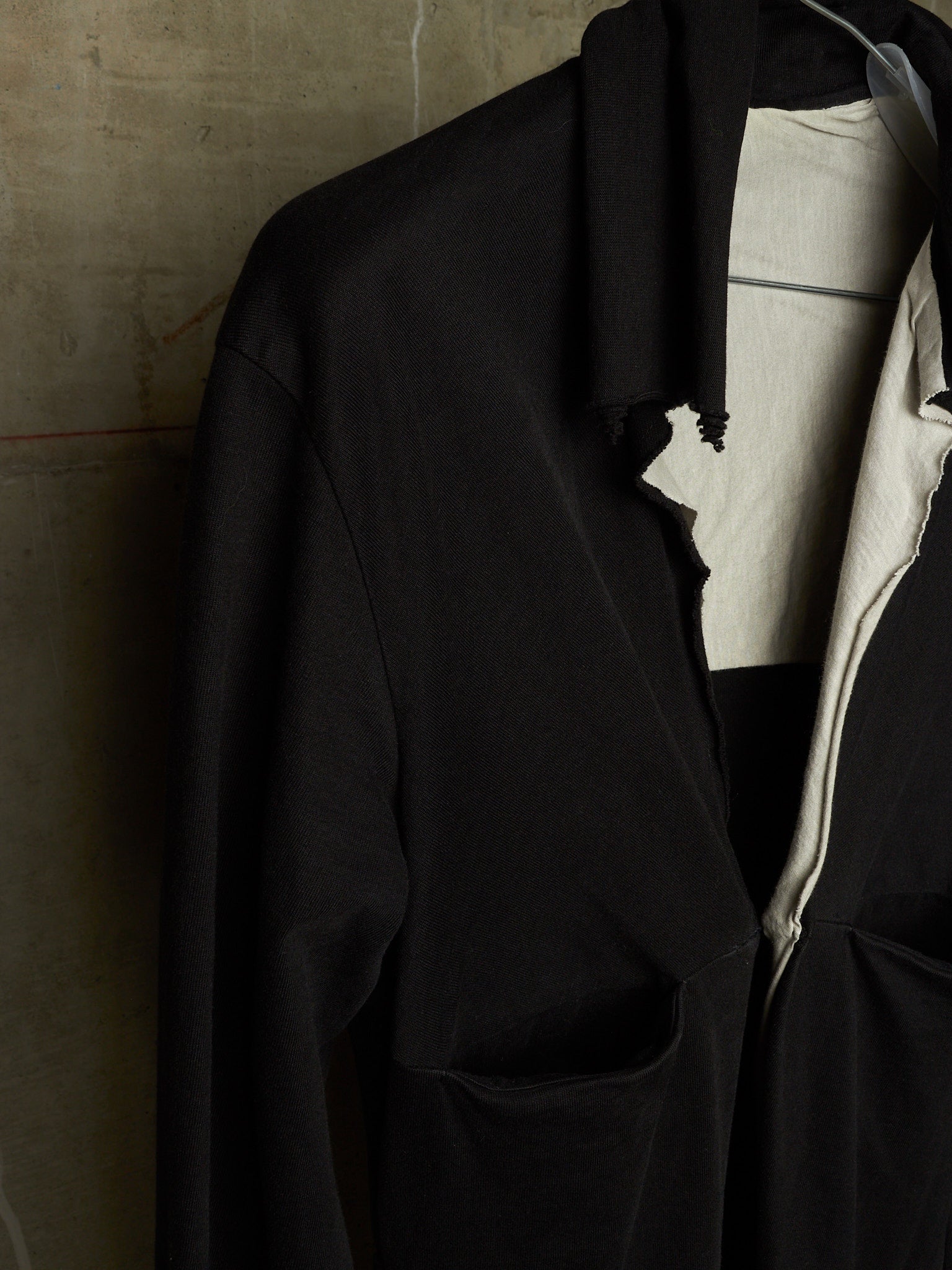 A black oversized jacket or overshirt with a raw, frayed collar and side slit, layered over a white shirt, shown against a textured concrete wall.