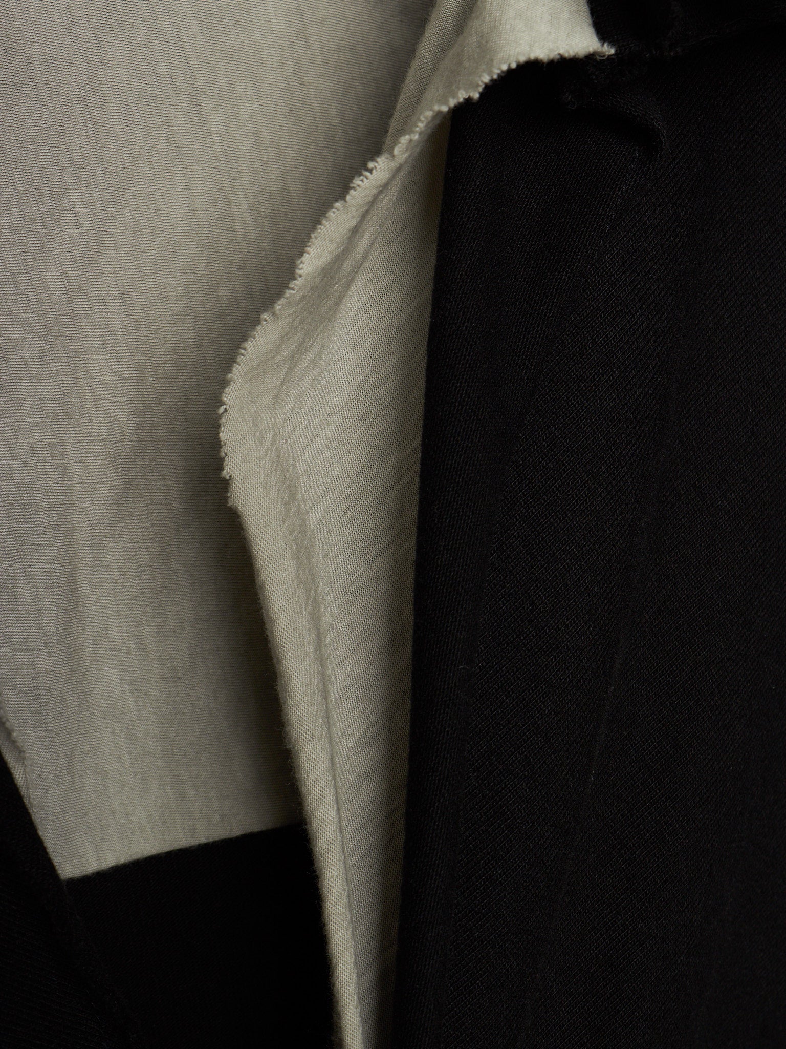Close-up of a garment showing two fabric panels: a light warm gray/beige textured panel on the left and a black panel on the right, with the beige fabric folded over to show its texture.