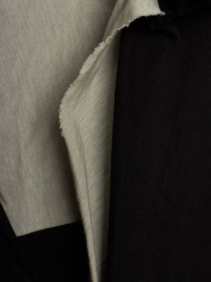 Close-up of a garment showing two fabric panels: a light warm gray/beige textured panel on the left and a black panel on the right, with the beige fabric folded over to show its texture.