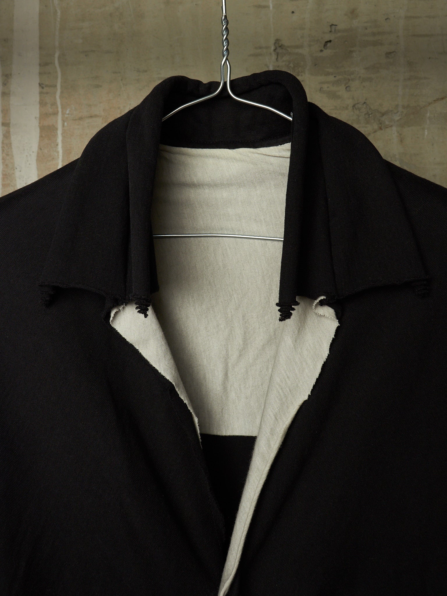 A black oversized coat or jacket with raw, frayed edges, layered over a light beige knit top and hanging on a wire hanger against a textured concrete wall.