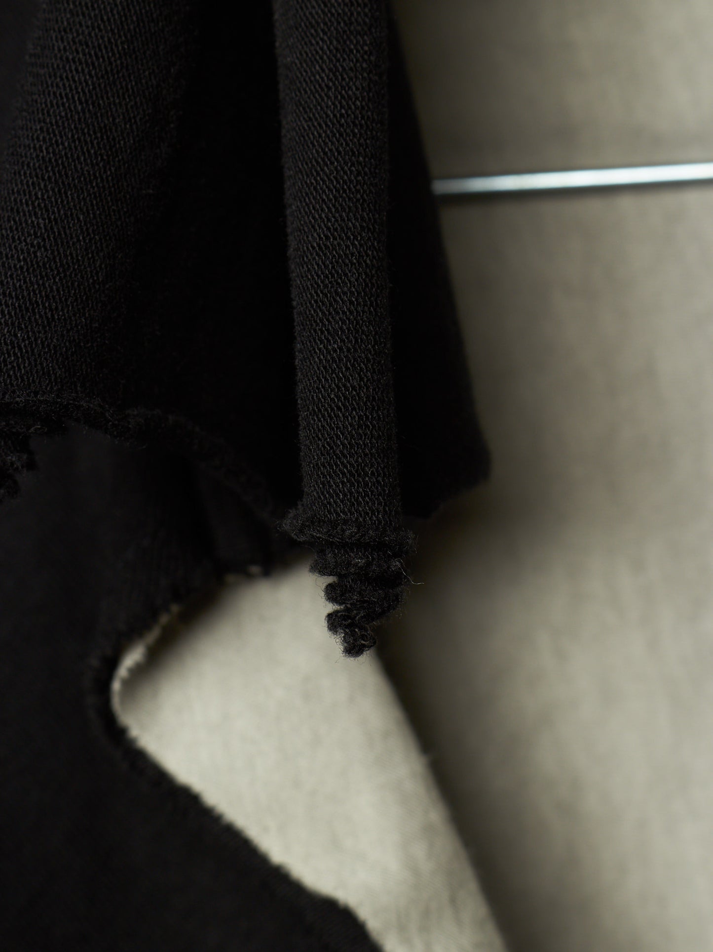 Close-up detail of a black knit garment with a frayed, cropped edge hanging against a neutral beige background.