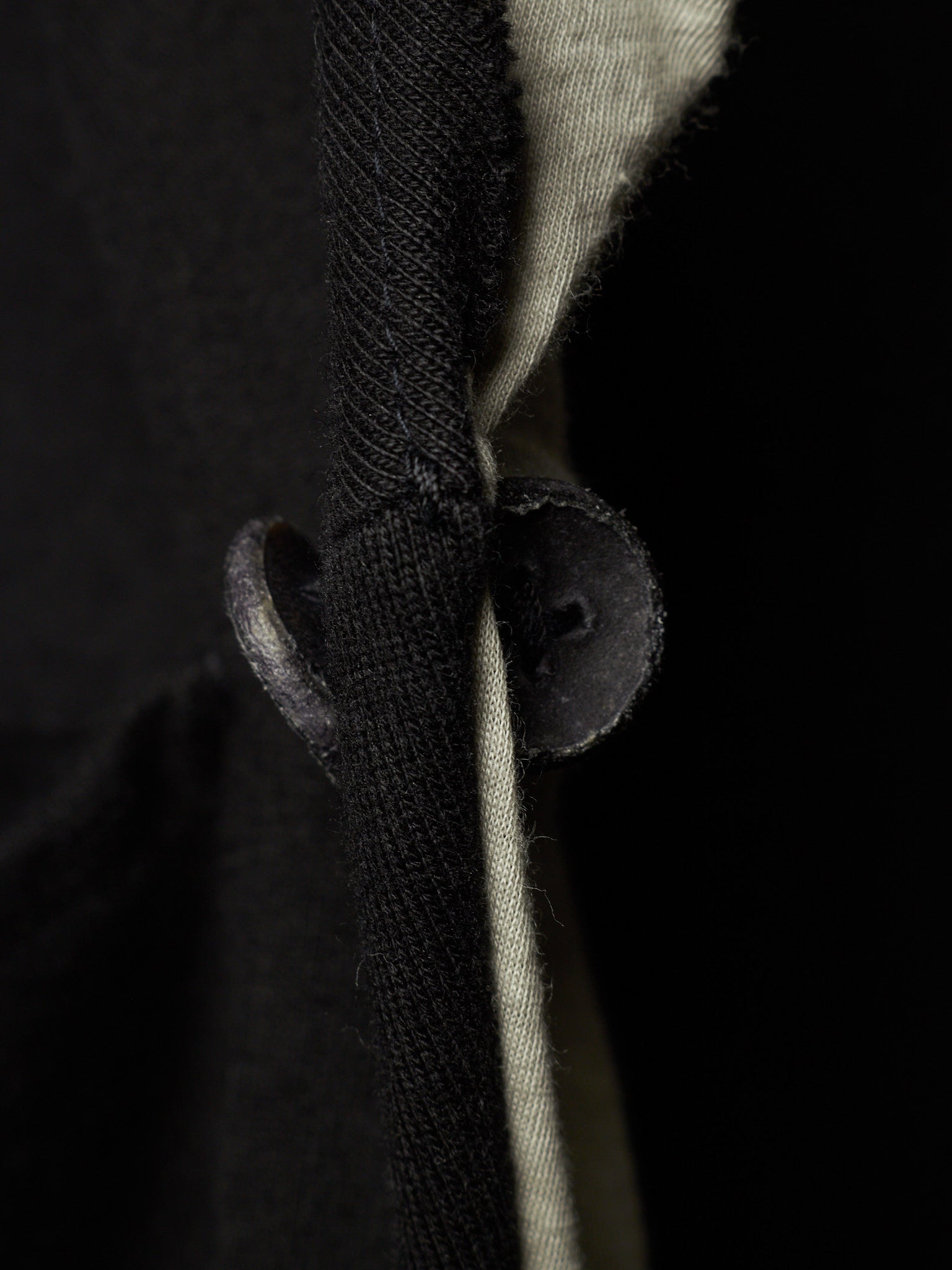 Close-up detail of a black knit garment with a dark button and a light beige inner lining or underlayer visible against a black background.