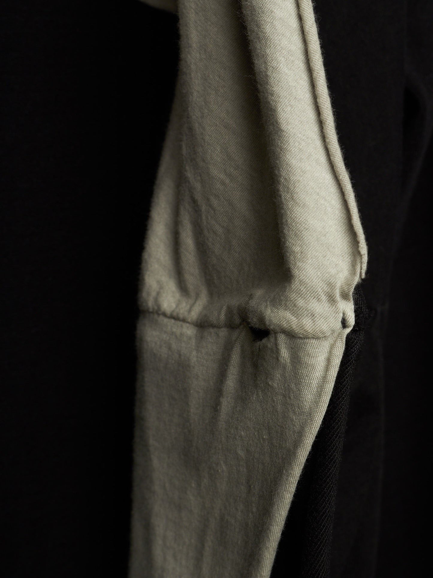 Close-up detail of a light beige or off-white fabric garment, showing a stitched seam and soft texture against a very dark black background.