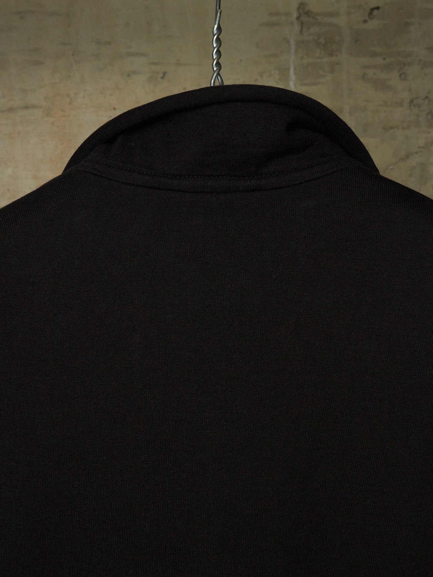 Close-up of the back of a black long-sleeve shirt hanging on a metal hanger against a textured concrete wall.