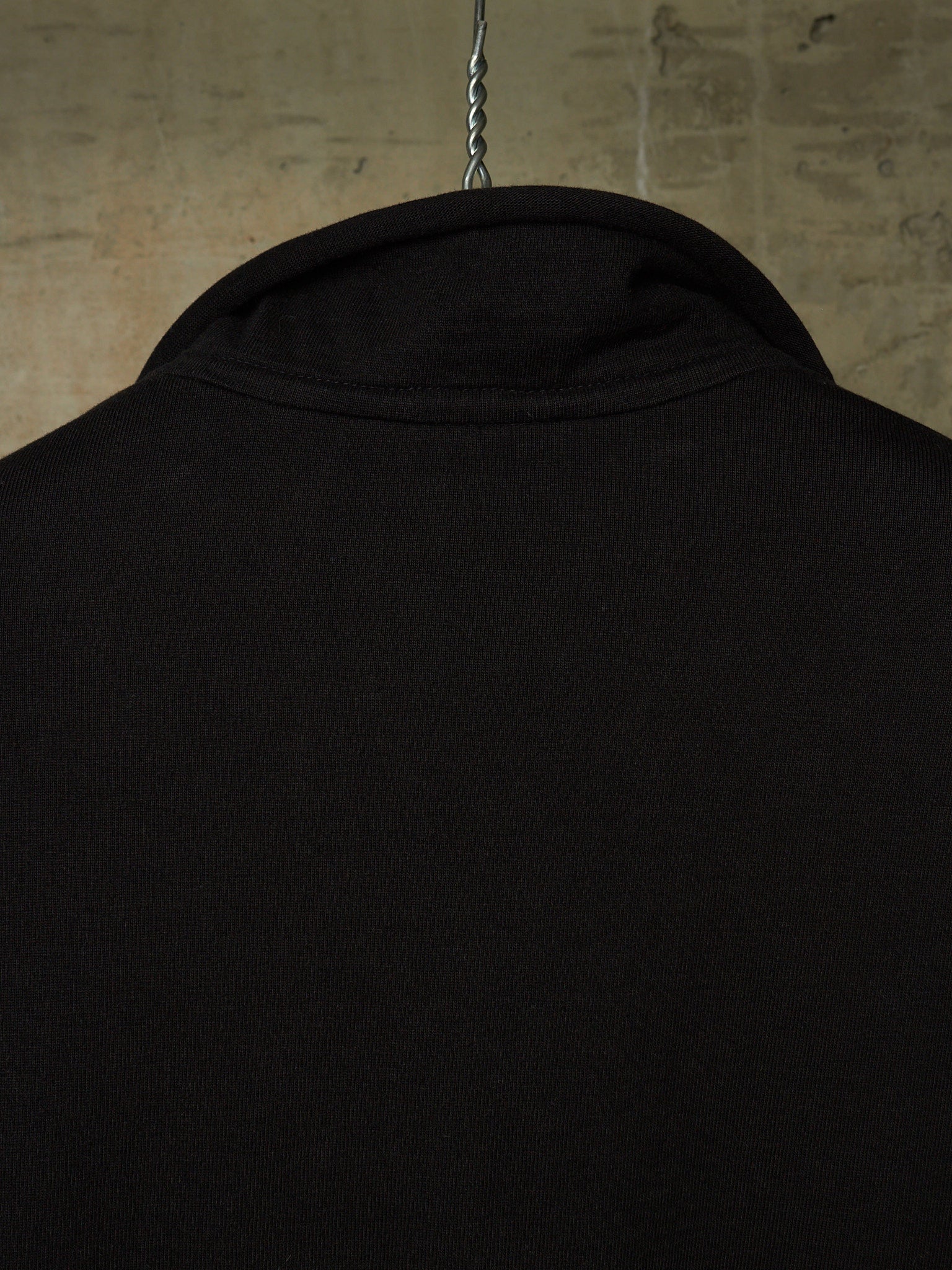 Close-up of the back of a black long-sleeve shirt hanging on a metal hanger against a textured concrete wall.