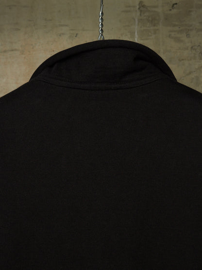 Close-up of the back of a black long-sleeve shirt hanging on a metal hanger against a textured concrete wall.