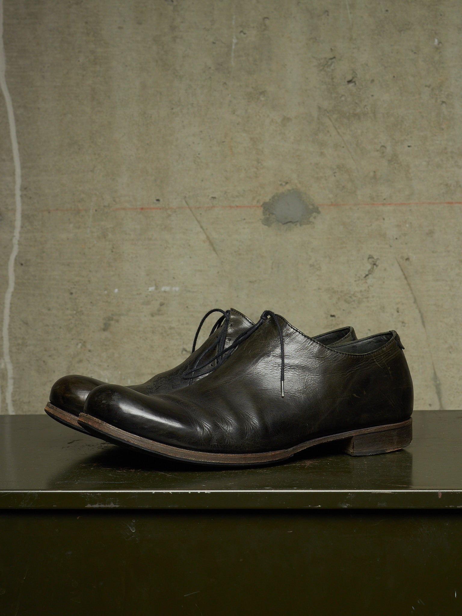 A pair of dark brown leather lace-up dress shoes resting on a dark green surface against a textured concrete wall.