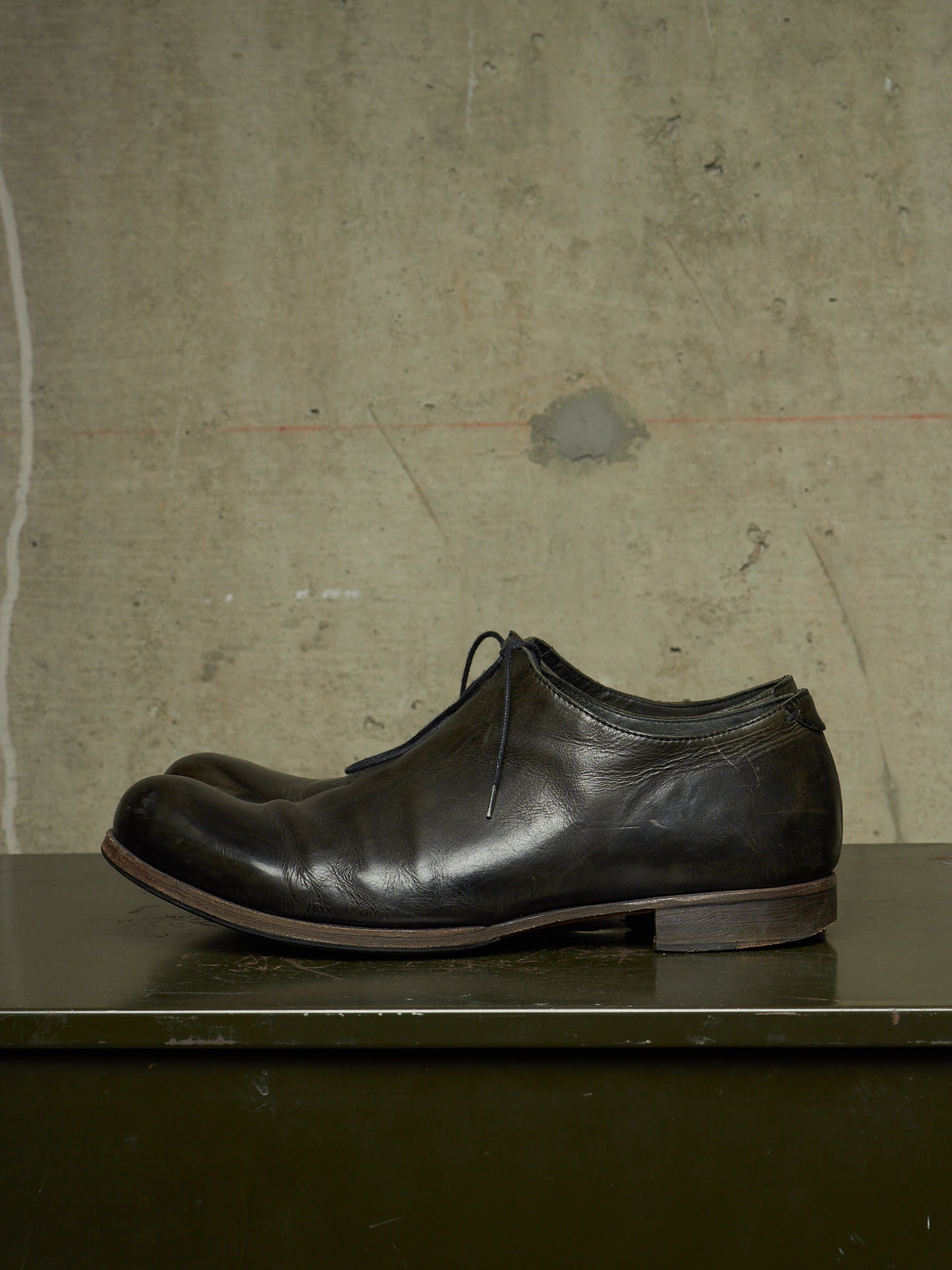 A pair of black leather lace-up dress shoes resting on a dark green surface against a textured concrete wall.