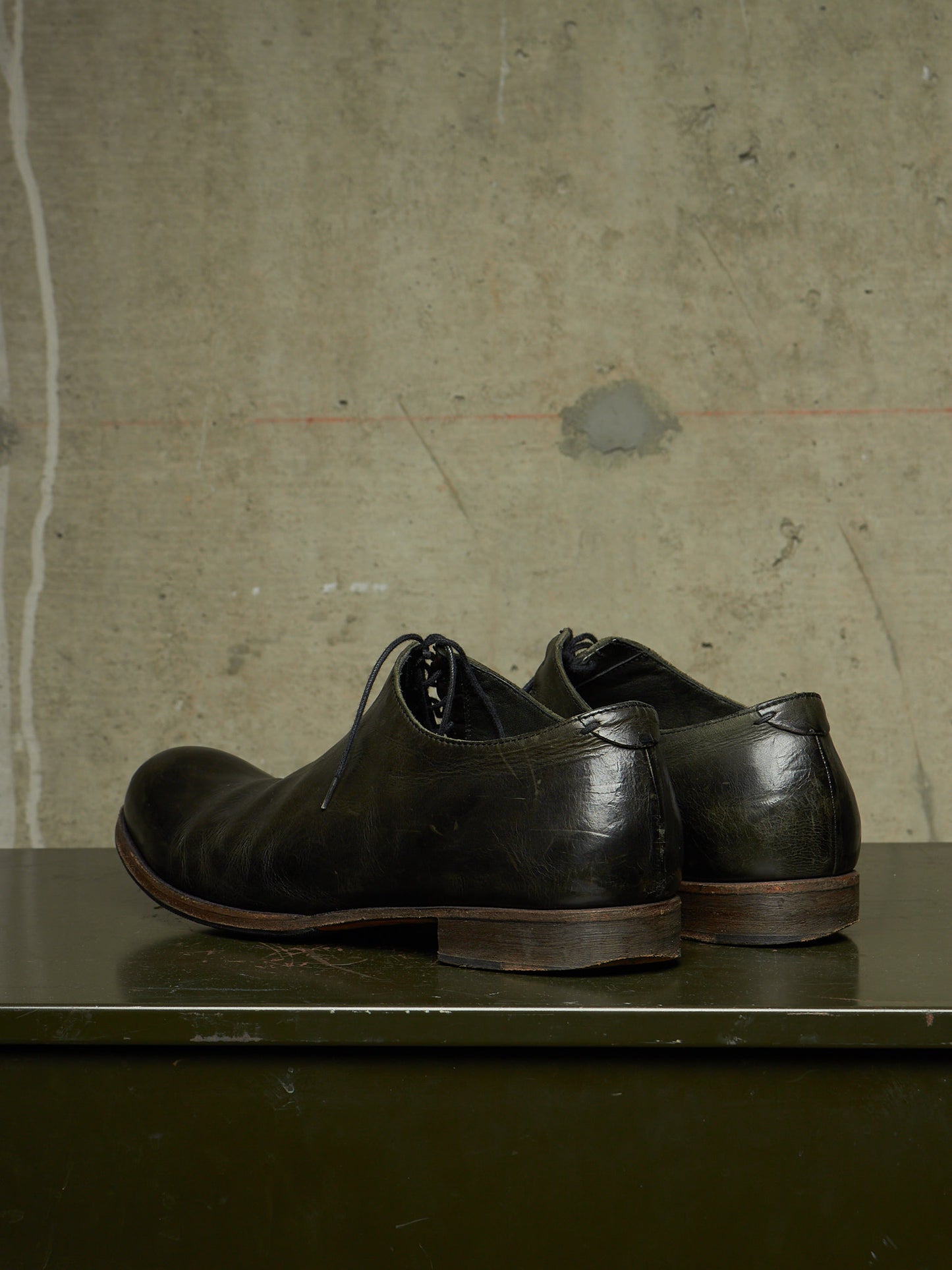 A pair of dark brown leather lace-up dress shoes with a distressed, vintage finish, displayed on a dark green surface against a textured concrete wall.