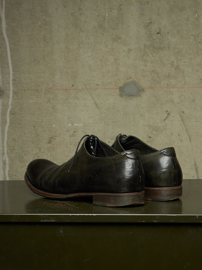 A pair of dark brown leather lace-up dress shoes with a distressed, vintage finish, displayed on a dark green surface against a textured concrete wall.